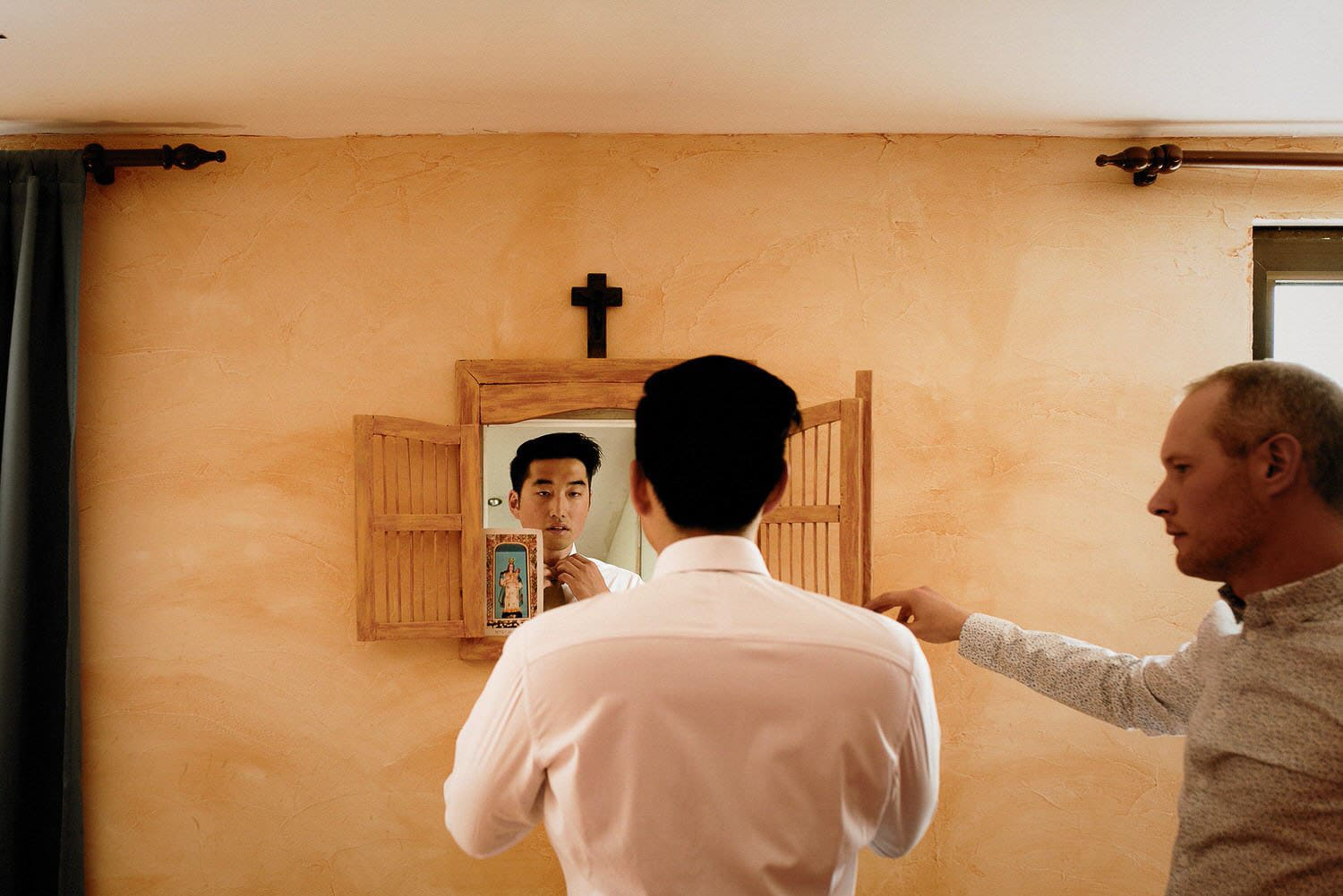 Charming Destination Wedding in the Portuguese Countryside - groom getting ready and looking in the mirror as he puts his tie on inside a rustic orange room